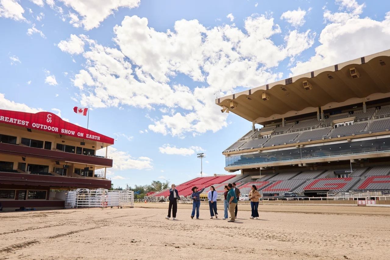 Group of people stand on rodeo arena dirt between grandstands under partly cloudy sky.
