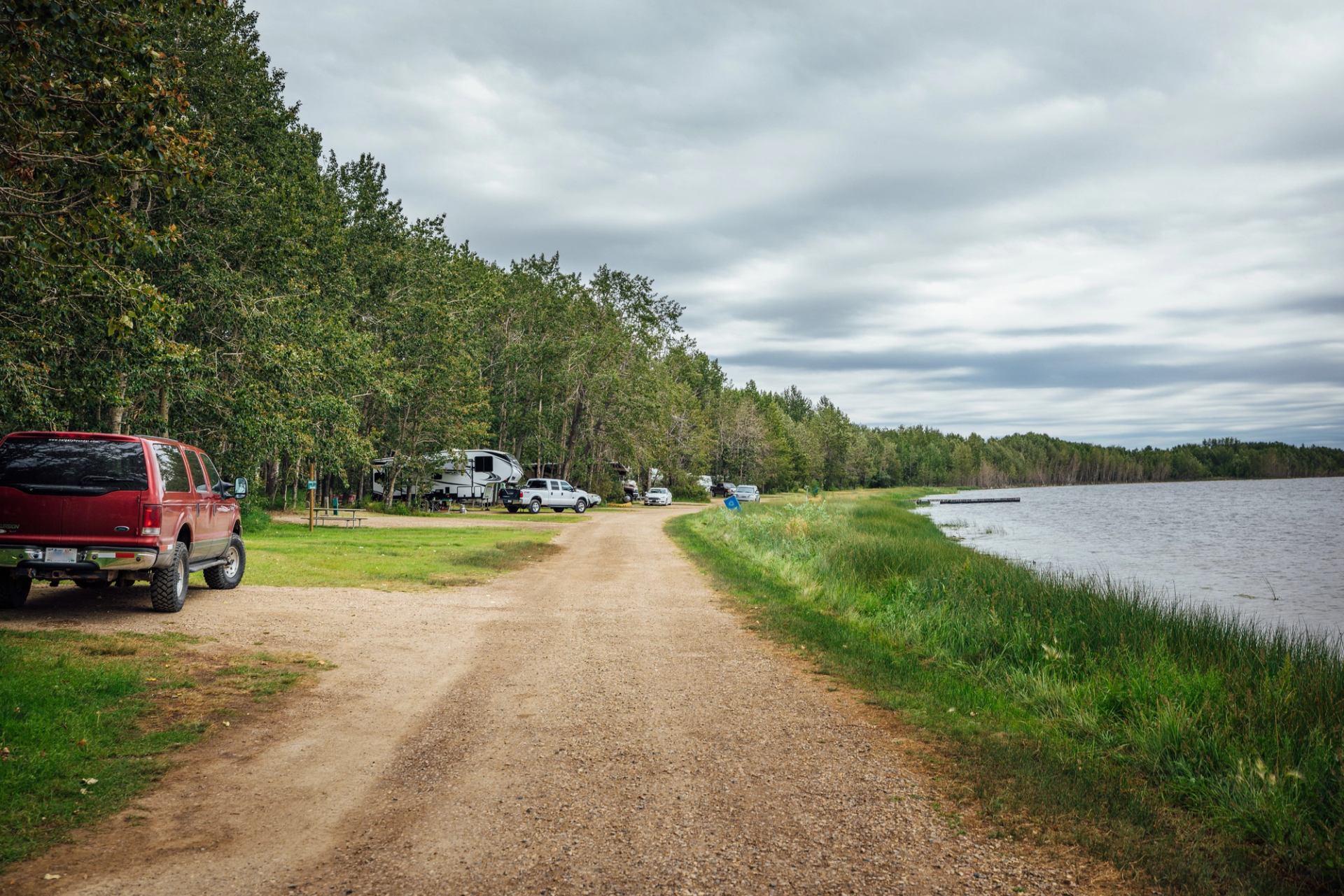 Gravel road along a lake with parked vehicles and RVs, surrounded by trees under a cloudy sky.