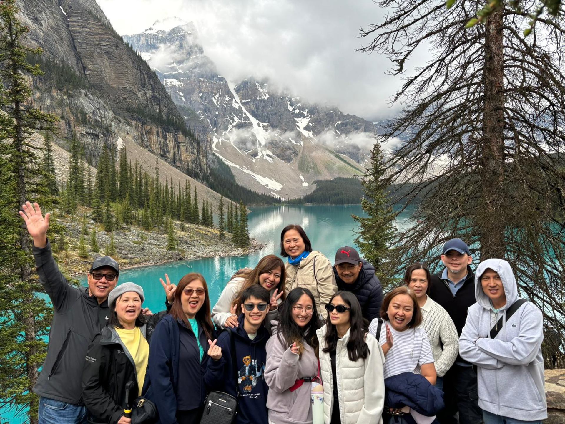 Group poses by turquoise lake with snow-capped mountains and forest backdrop.