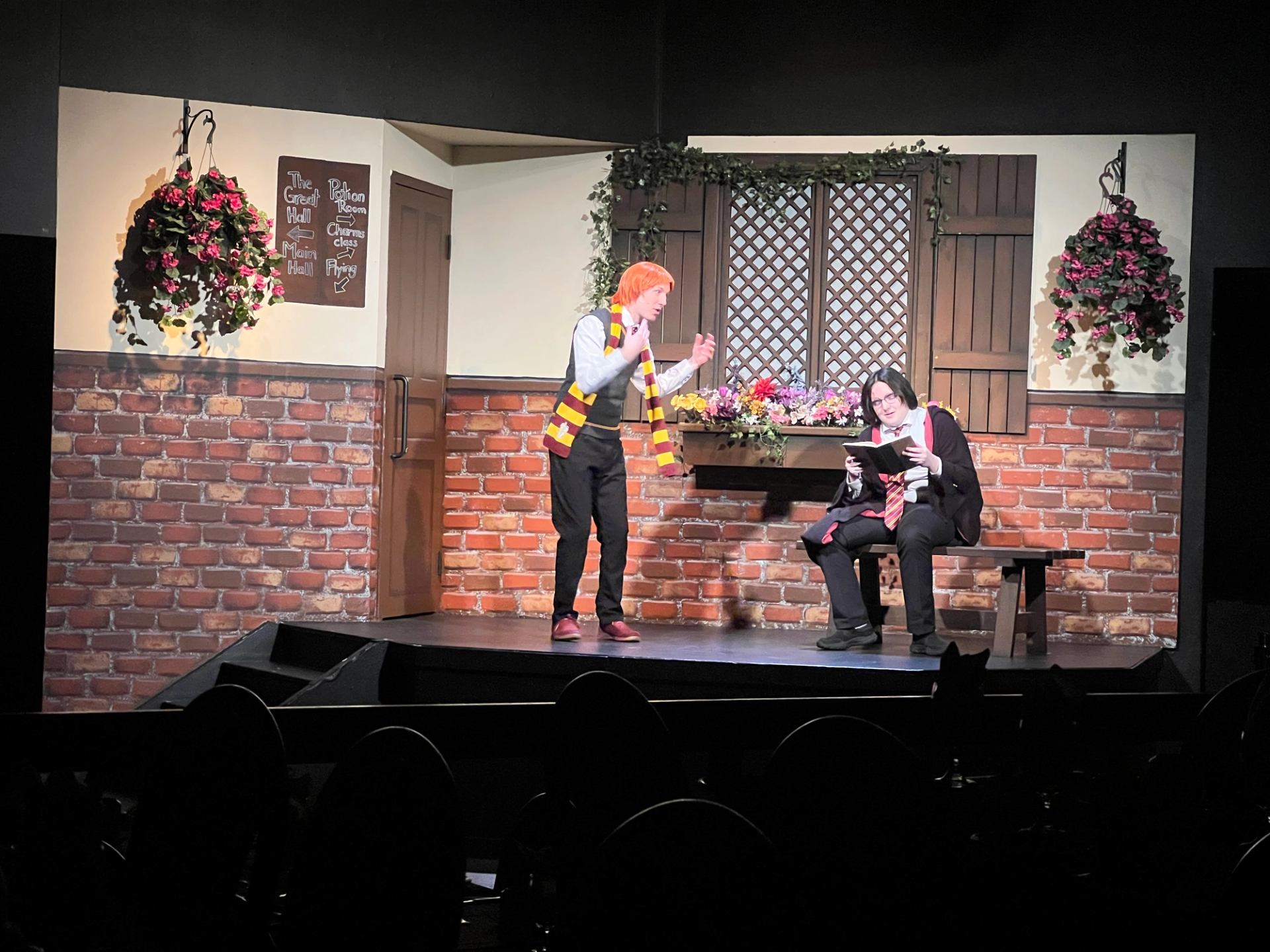 Stage performance with two costumed actors, one standing and one seated reading.
