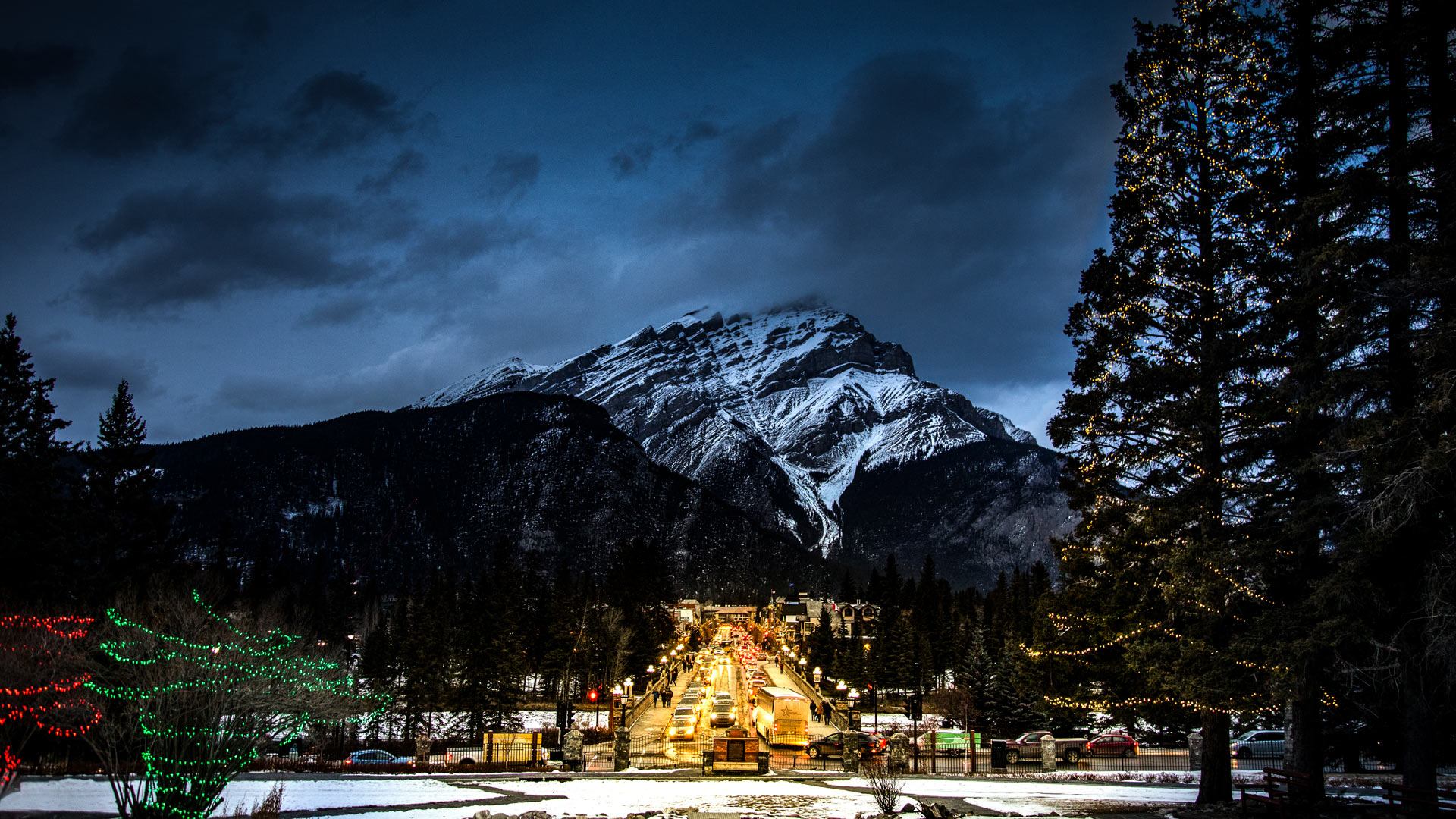 Mountain peak with snow-covered slopes above a lit town street and trees at dusk.