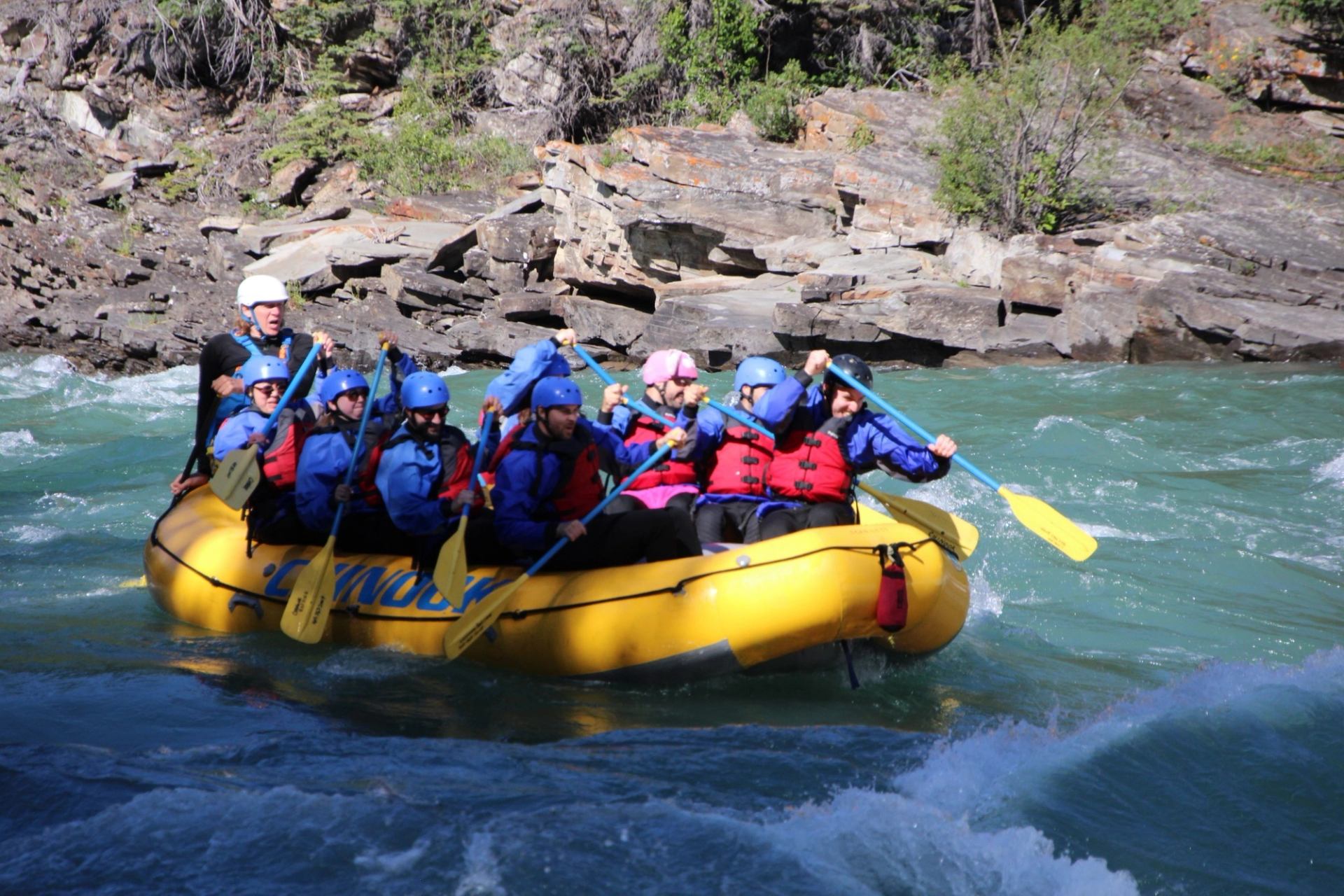 Group in helmets and lifejackets paddles yellow raft through turquoise river rapids.