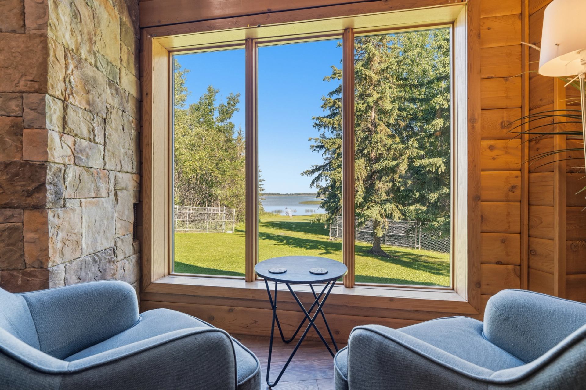 Lakeside view through large cabin window with two blue chairs and round table.