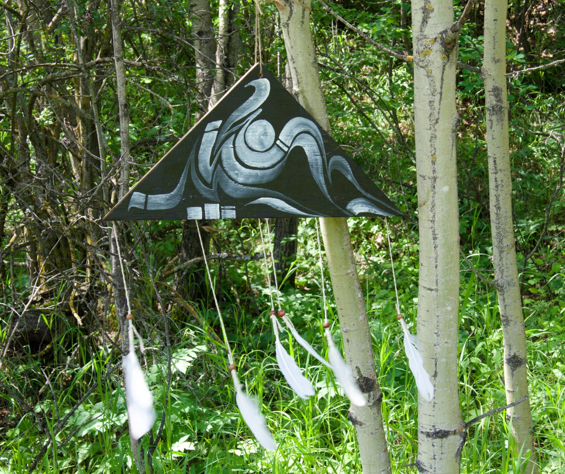 Triangular wooden artwork with abstract design and hanging white feathers among trees.