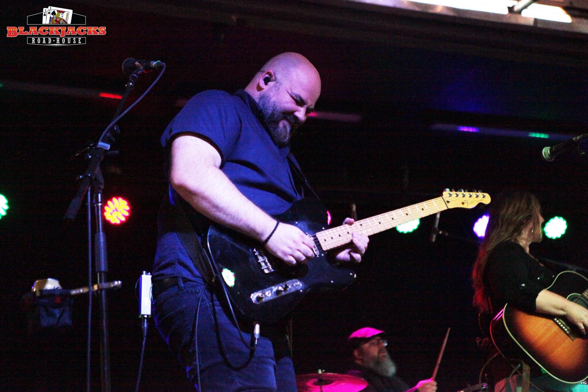 Guitarist performs on stage, singing into a microphone at Blackjack’s Roadhouse concert.