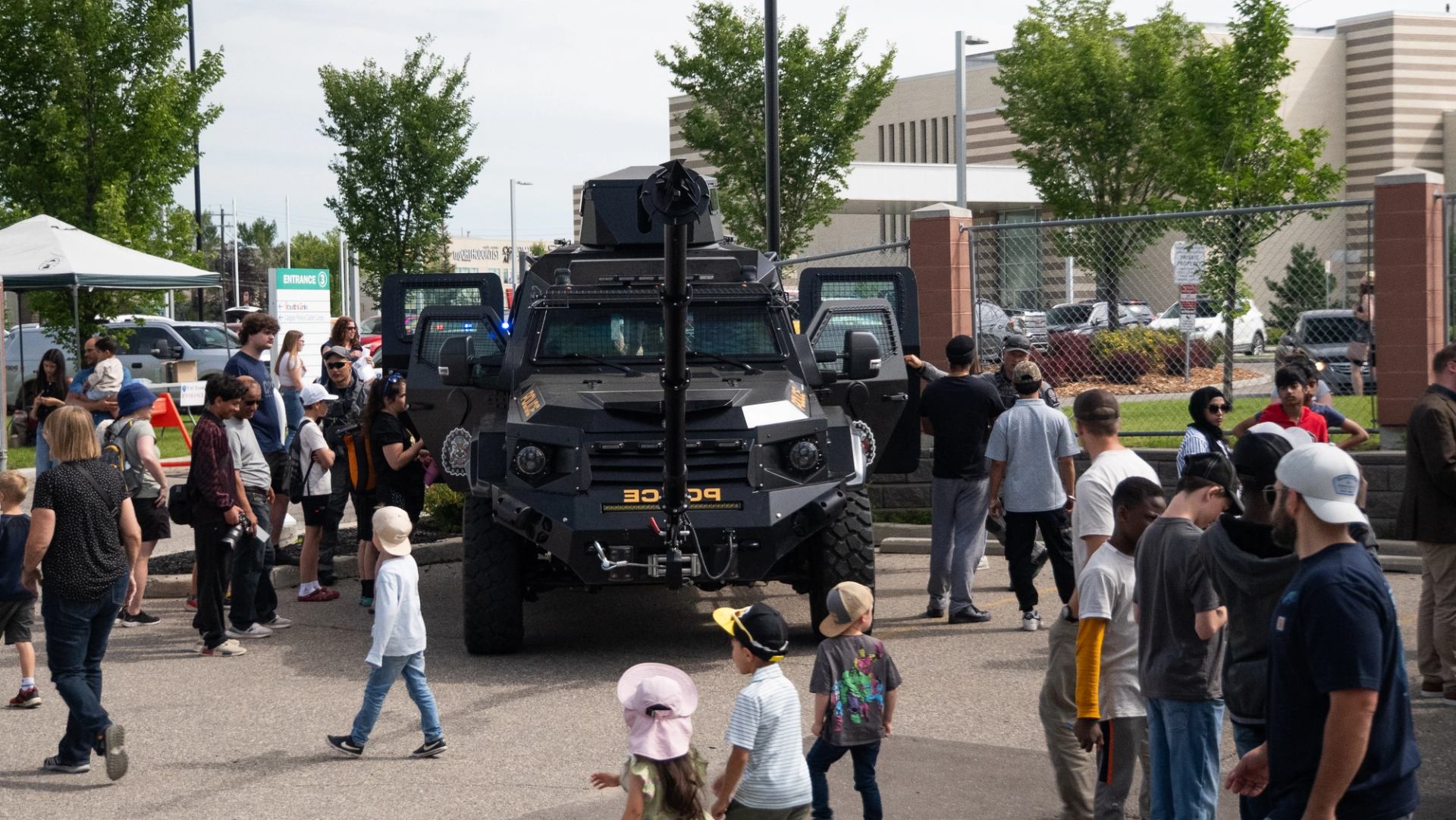 Crowd gathers around a black armoured police vehicle with open doors.