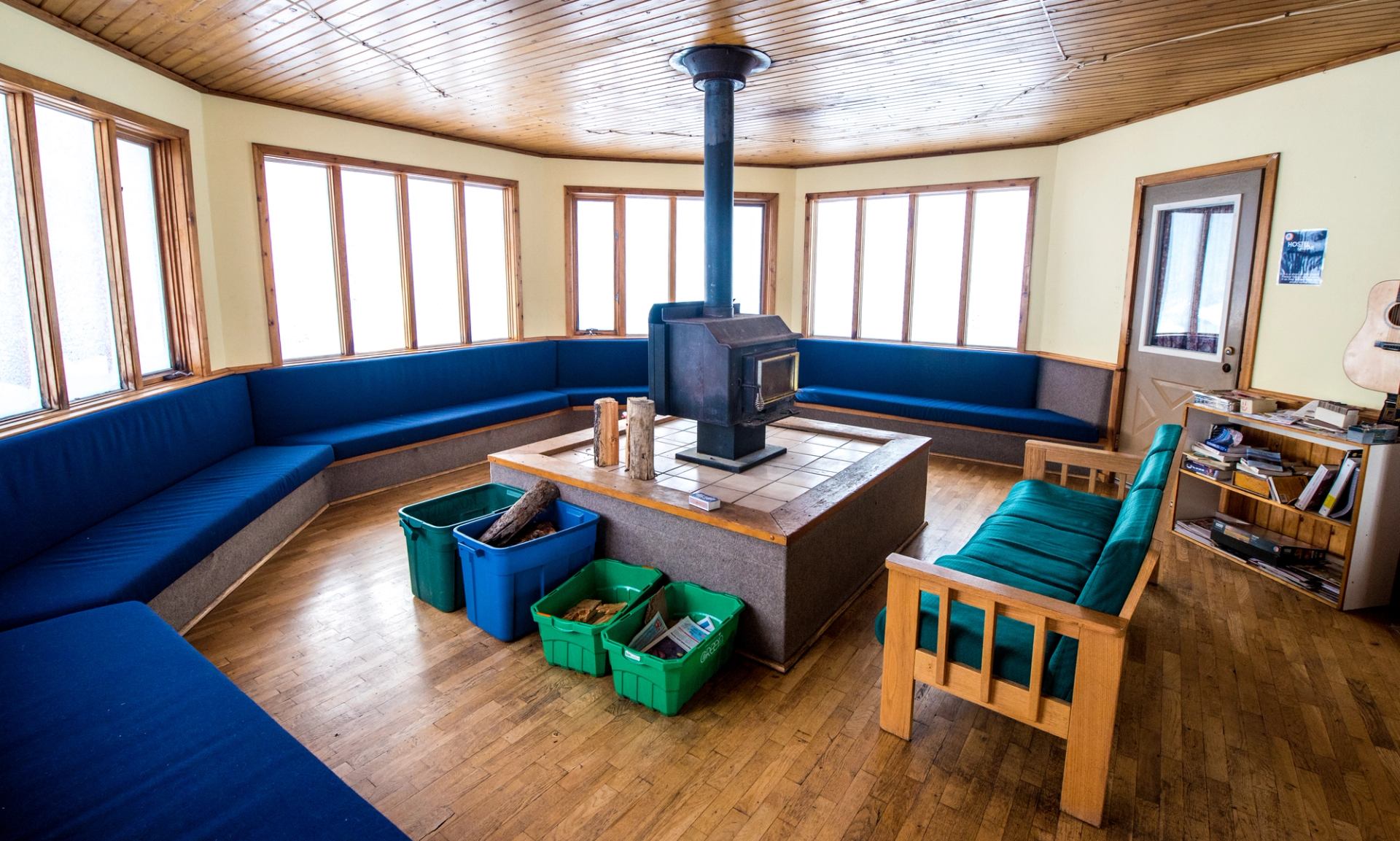 Common area with wraparound windows, cushioned bench seating, and central wood stove.