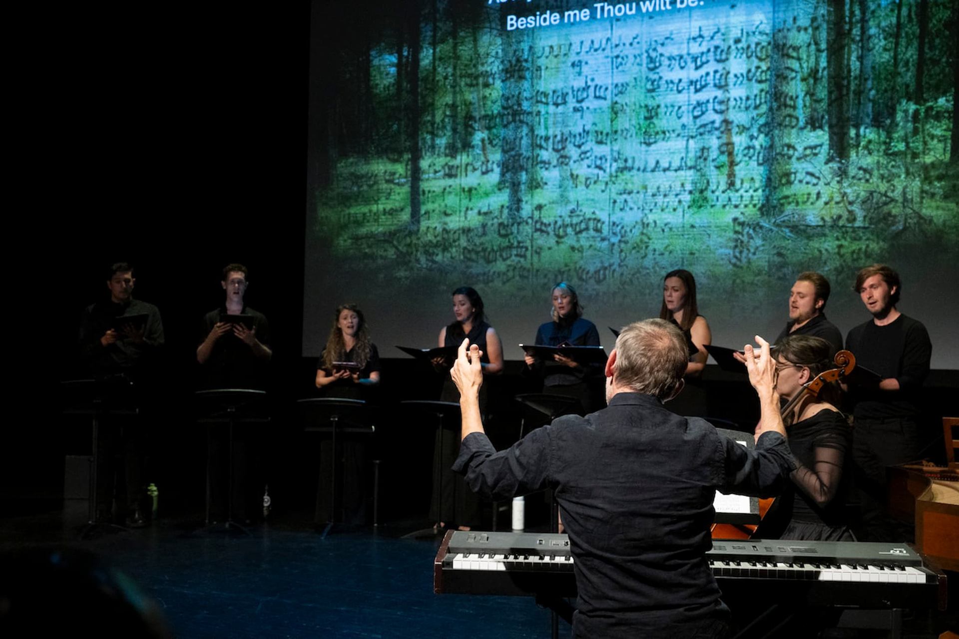 Conductor leads choir and musicians onstage before large screen with forest and music.