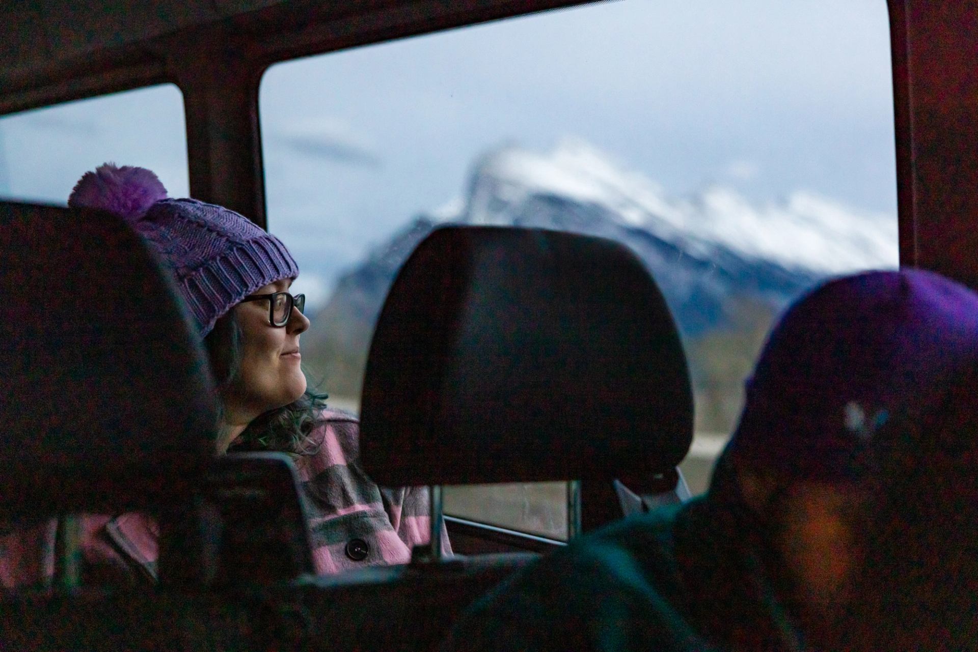 Person in purple toque looks out vehicle window at snow-covered mountain landscape.