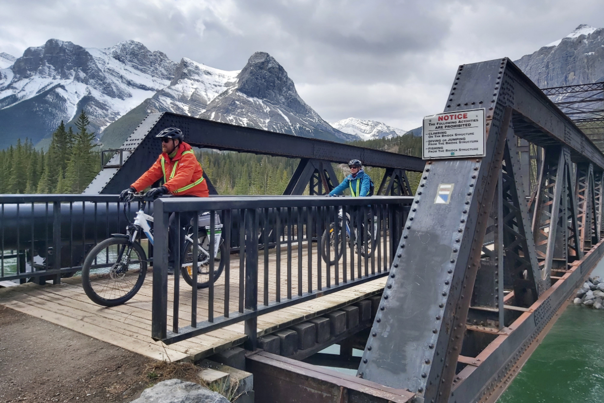 Cyclists ride across a metal bridge over green water with snow-covered mountains behind..