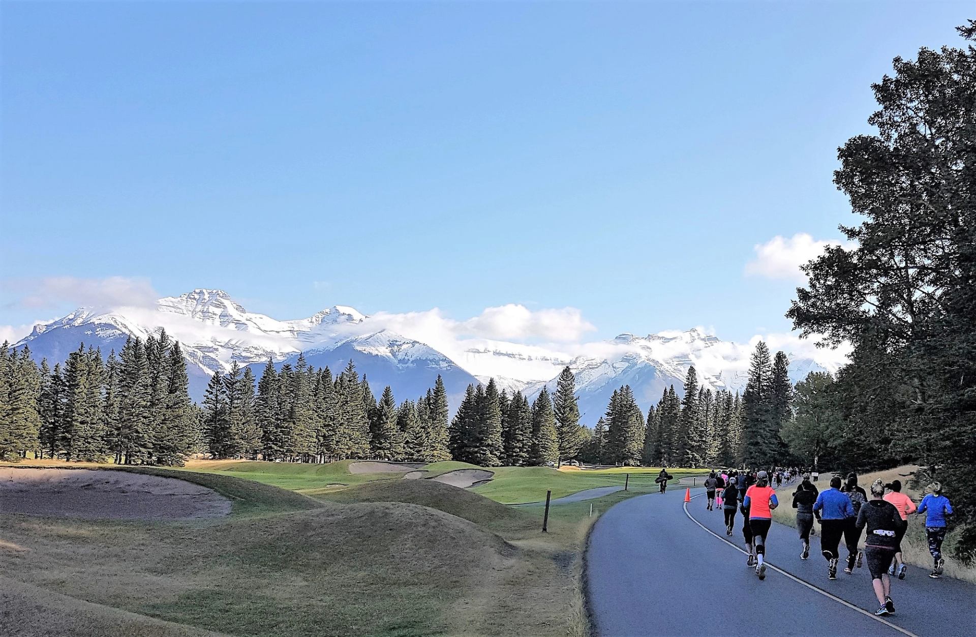 Runners on a paved path next to a golf course, with snow-capped mountains and pine trees in the background.