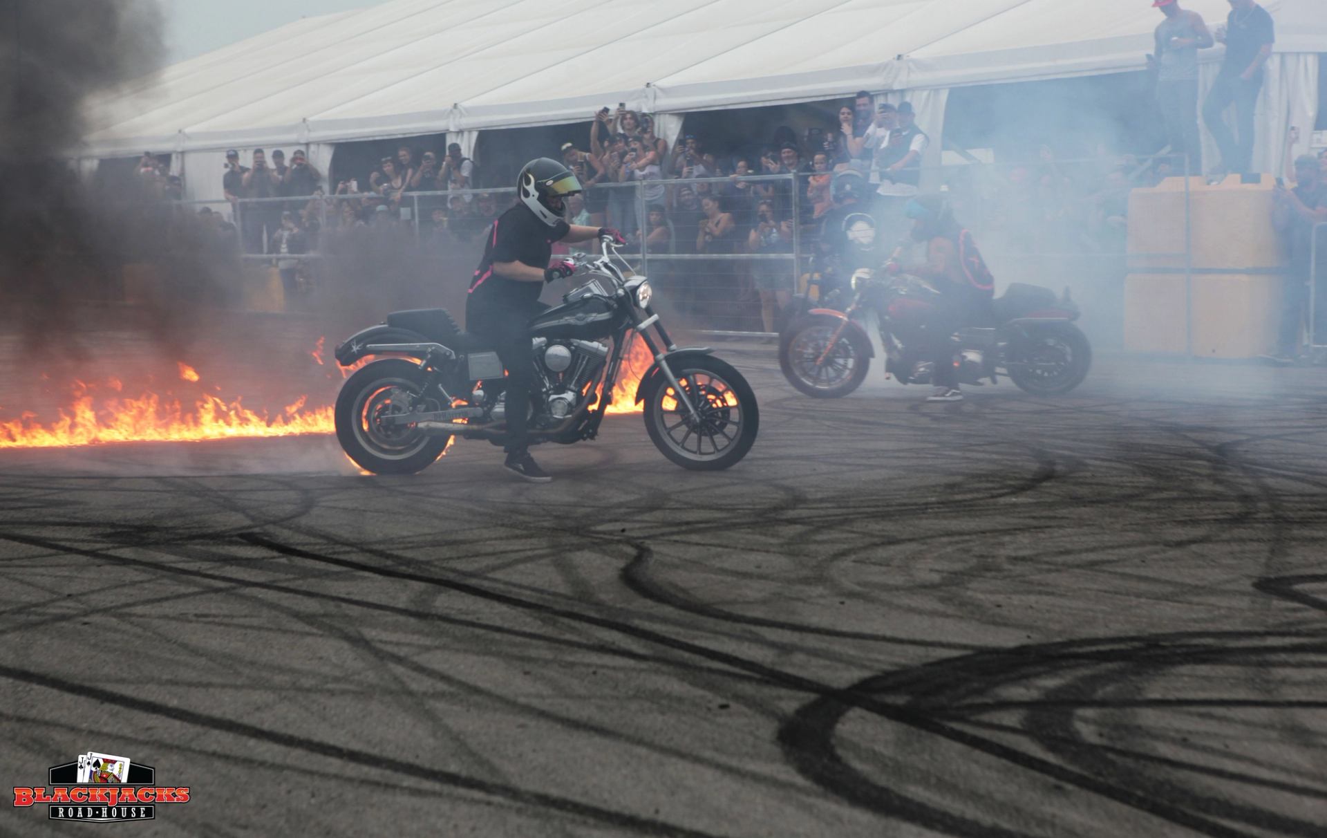 Two motorcyclists perform stunts in front of a crowd at Blackjacks Roadhouse. One rider is surrounded by flames and smoke as tire marks cover the pavement, while spectators watch from behind a fence near a large white event tent.