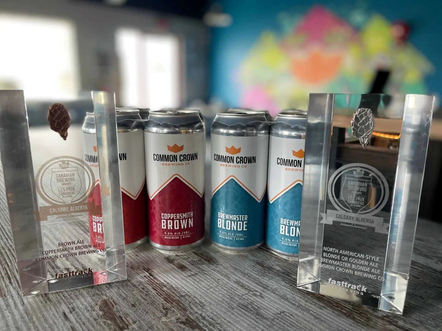 Three Common Crown beer cans with four awards on a wooden surface.