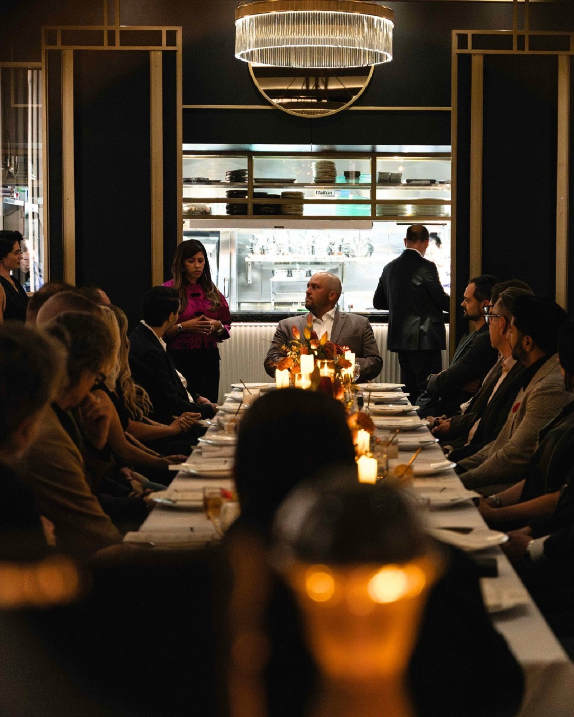 Long candlelit dinner table with people seated, listening to a speaker in a restaurant.