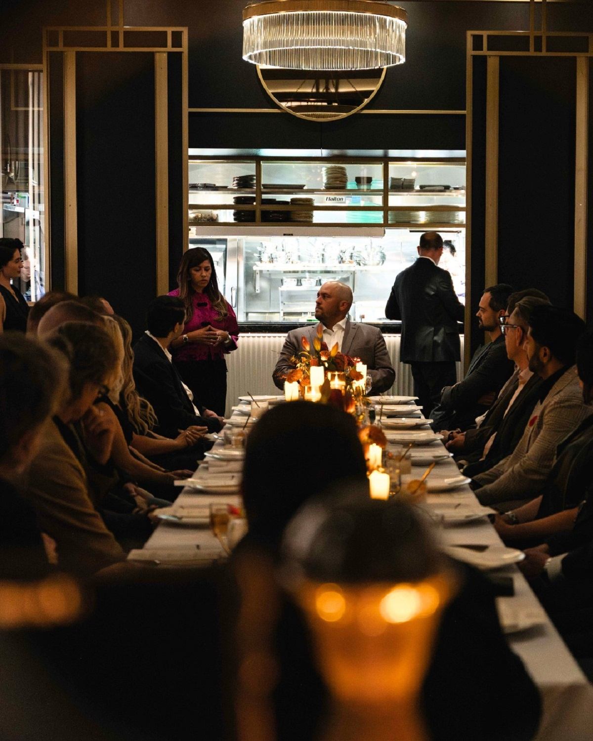 Long candlelit dinner table with people seated, listening to a speaker in a restaurant.