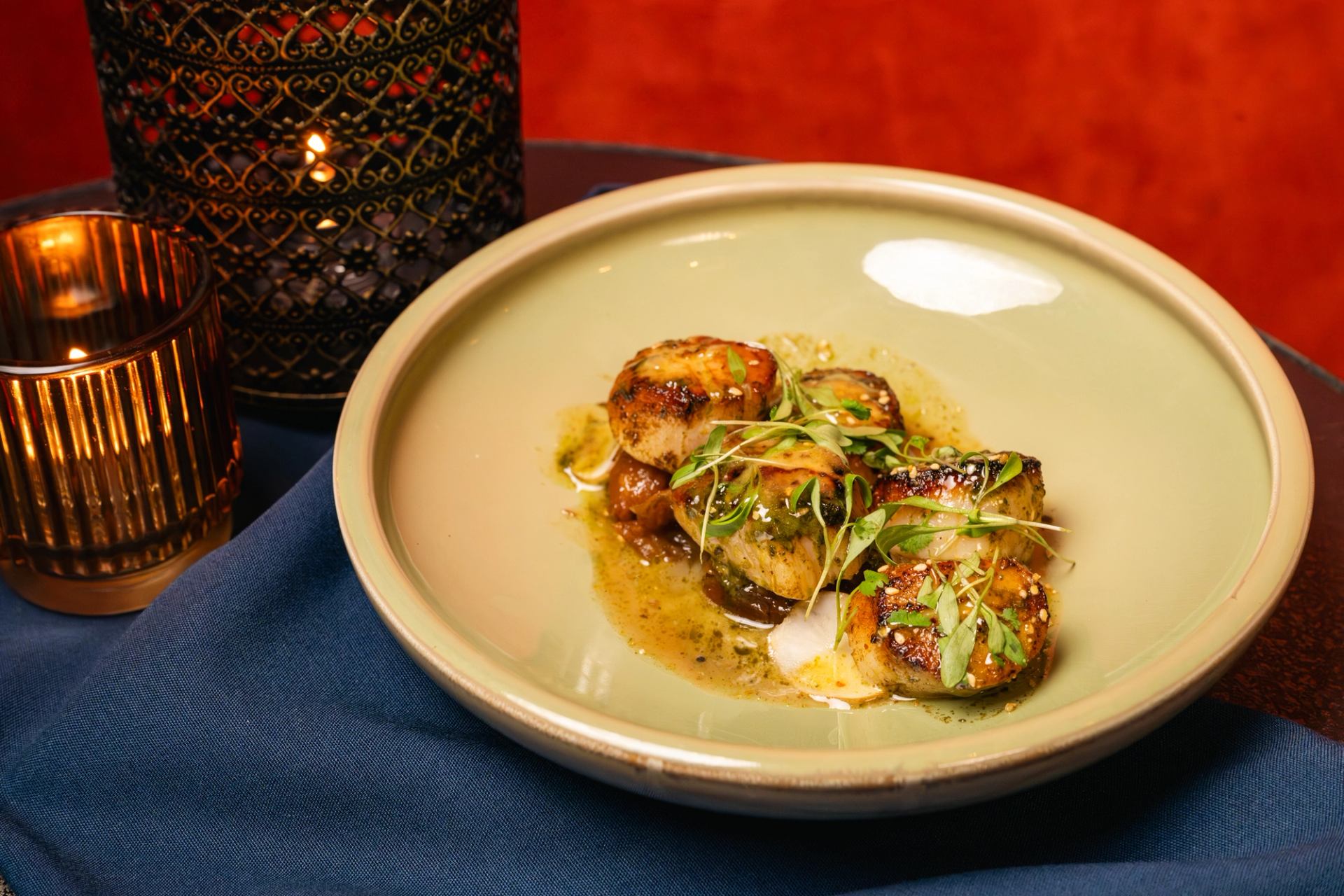 Seared scallops with sauce and microgreens on beige plate beside lit candles.