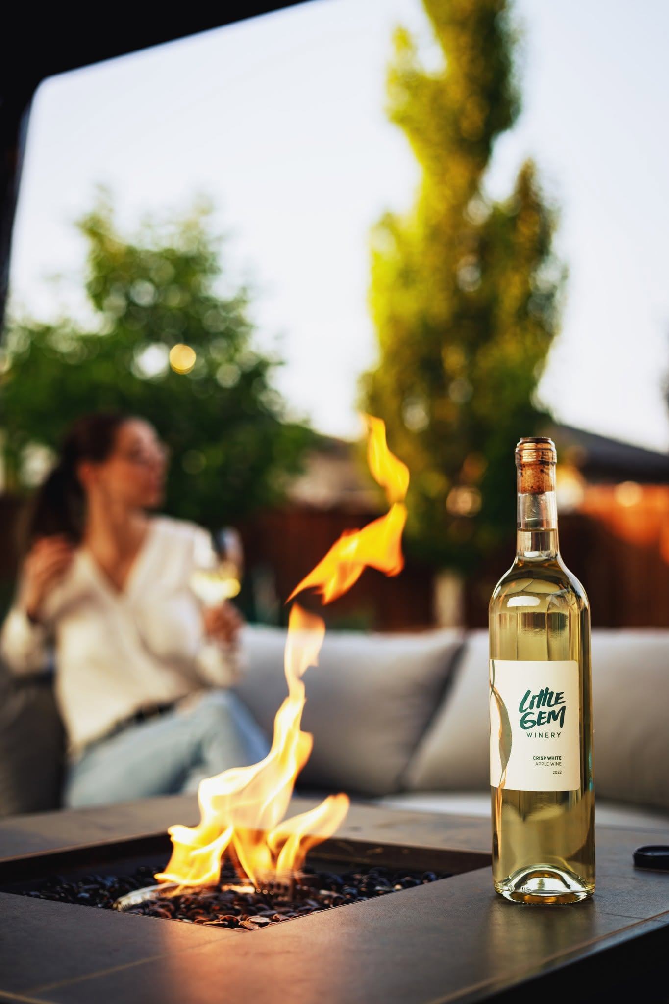 Bottle of Little Gem white wine beside gas firepit, person holding glass in background.