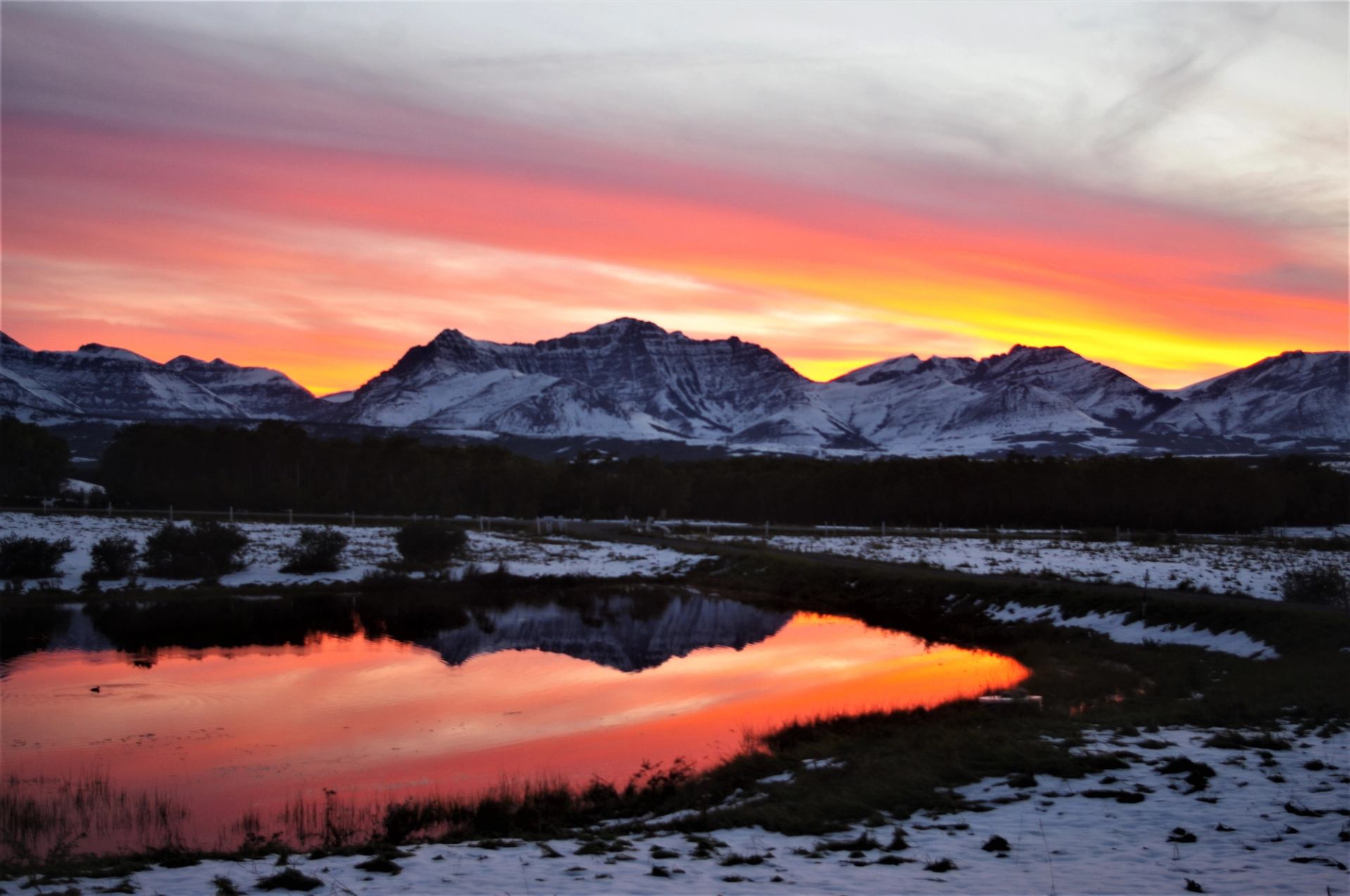 Snow-covered mountains beneath a red-orange sky reflected in a pond.