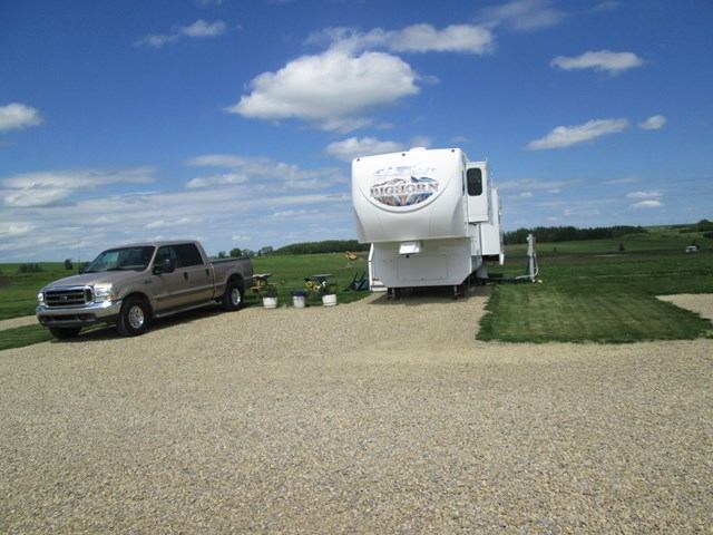 RV trailer and pickup truck parked on gravel beside grassy field with golf-course scenery.