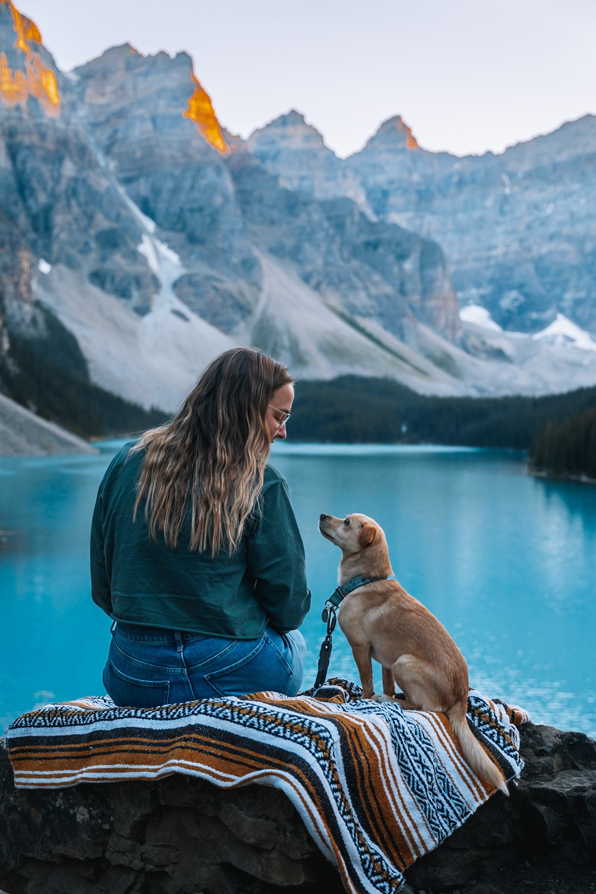 A woman and her small dog sitting on a blanket overlooking the blue water of Moraine Lake at sunrise