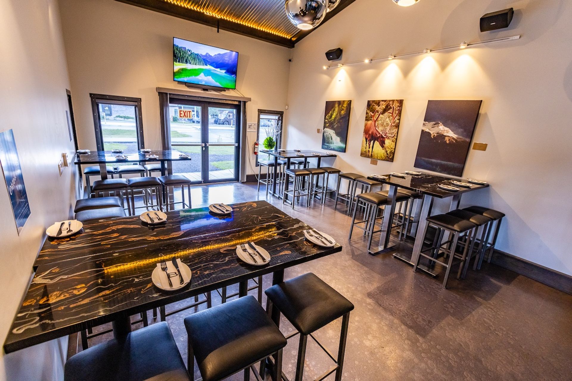 A modern restaurant interior with black and gold high-top tables, matching stools, nature-themed artwork on the walls, and a mounted TV screen.