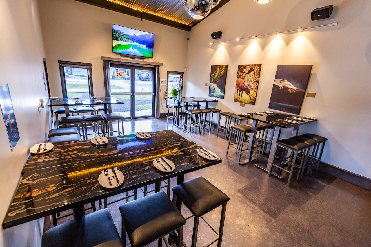 A modern restaurant interior with black and gold high-top tables, matching stools, nature-themed artwork on the walls, and a mounted TV screen.