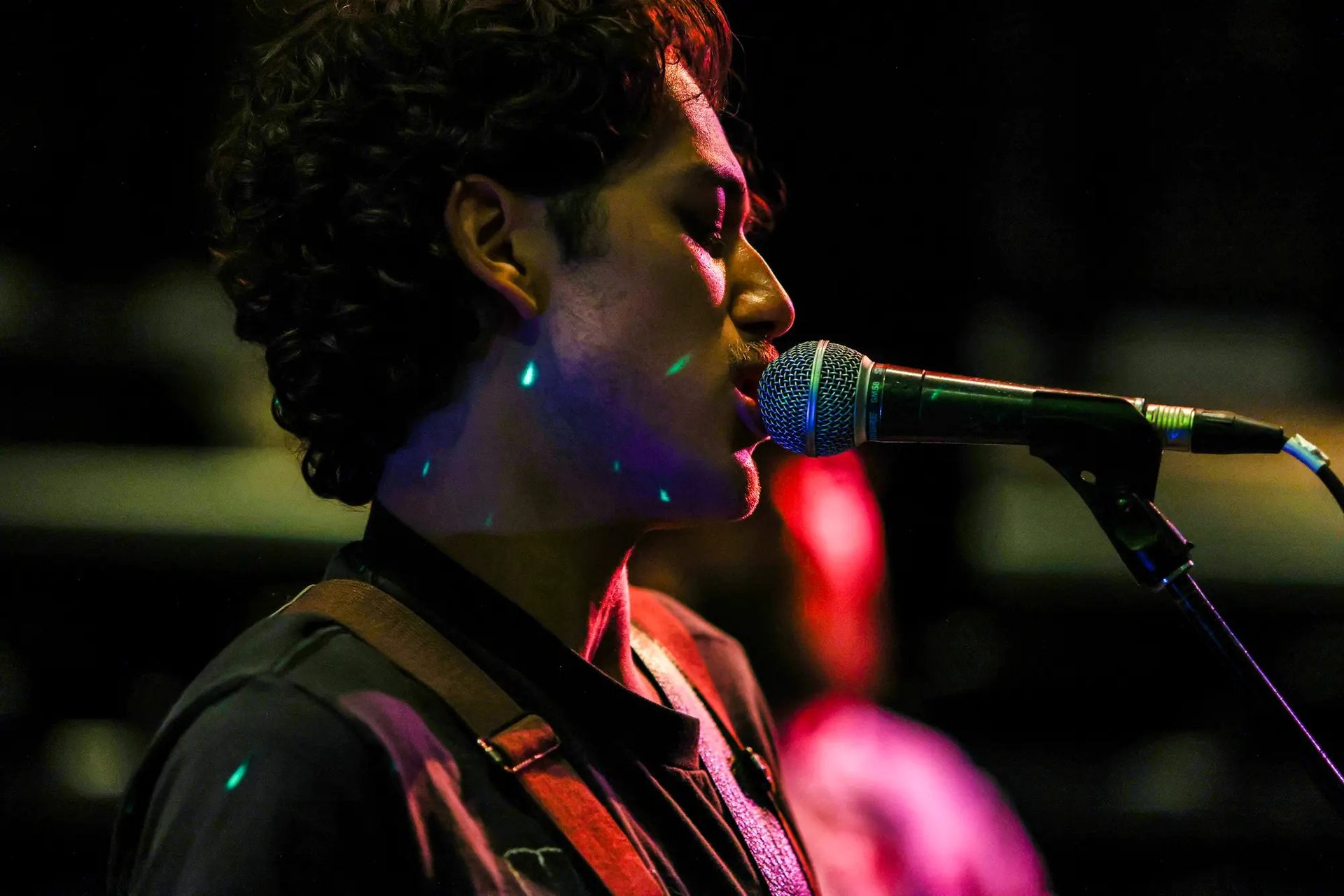 Musician sings into a microphone under colourful stage lights, wearing a guitar strap.