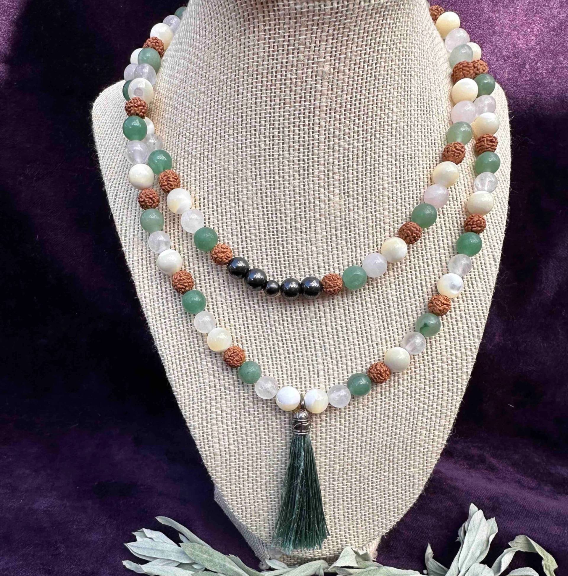 Beaded gemstone mala necklace with green tassel displayed on burlap bust against purple fabric.