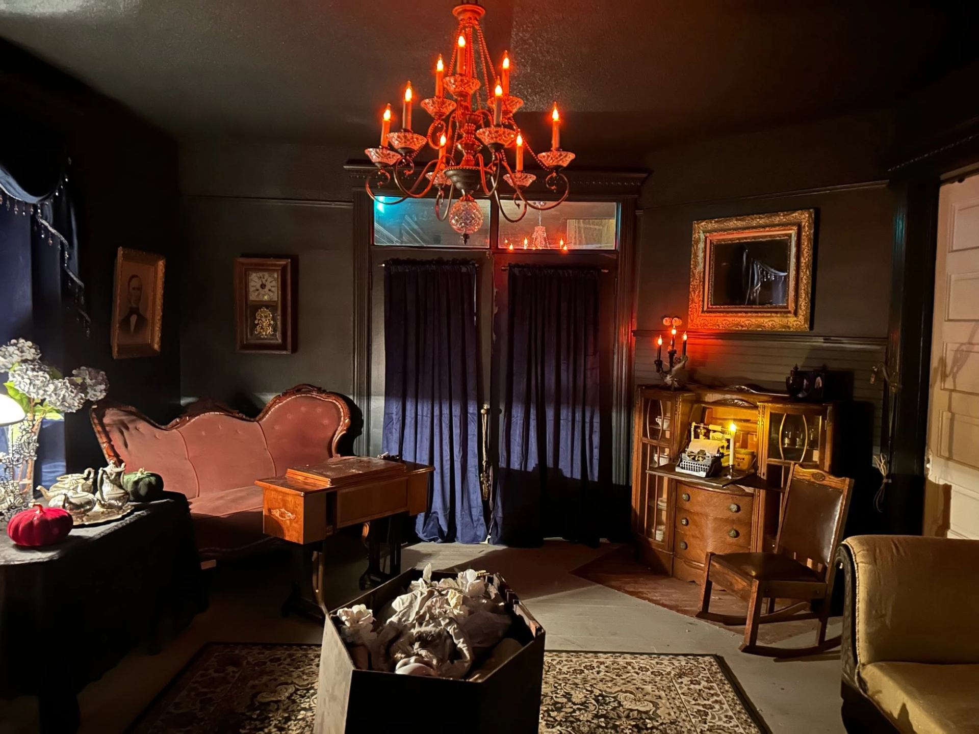 Dim Victorian-style parlour with red chandelier, antique furniture, typewriter and dark curtains.