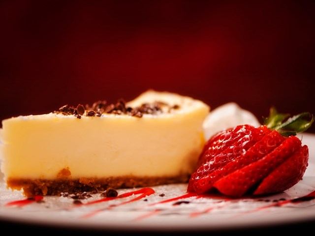 Slice of cheesecake with chocolate shavings, strawberry and red sauce on plate.