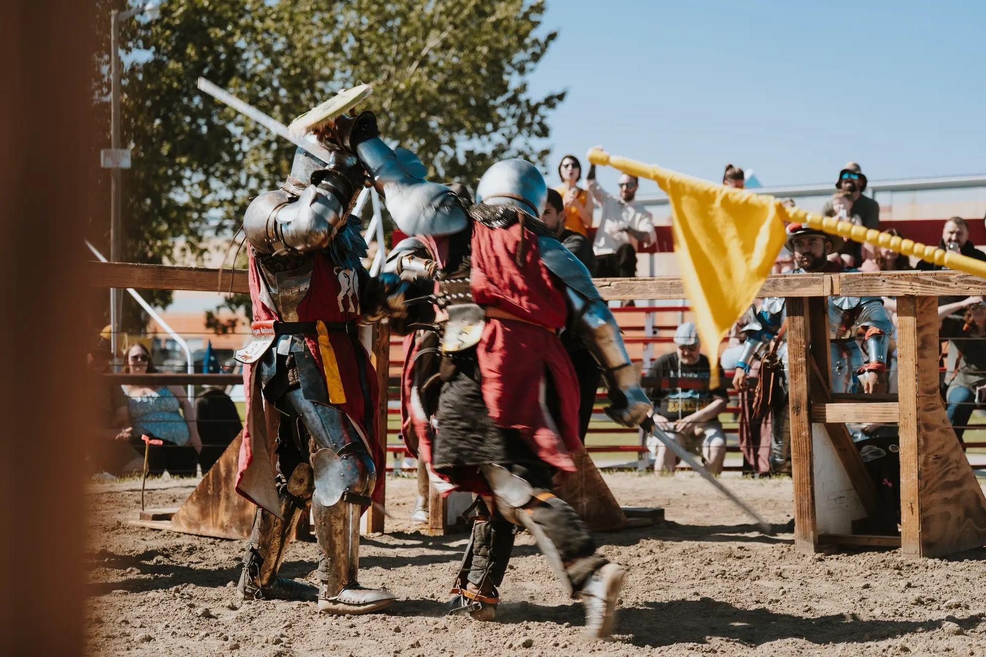 Armored fighters duel with swords at outdoor medieval faire with spectators and flags.