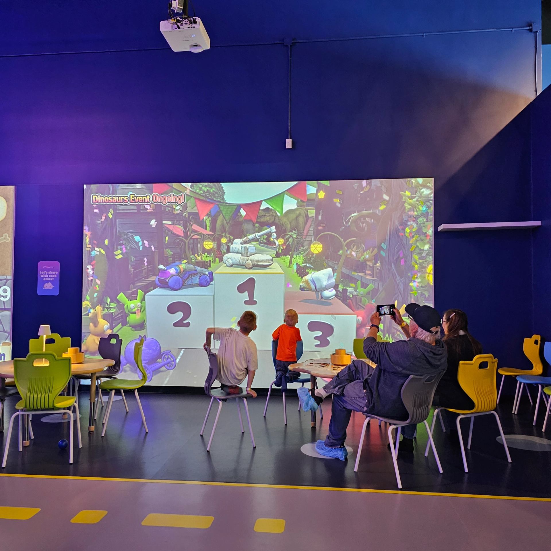 Kids and adults interact with a colorful projection game in a playroom with bright chairs.