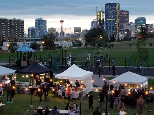 Outdoor market tents with string lights in park, Calgary Tower and skyline beyond.
