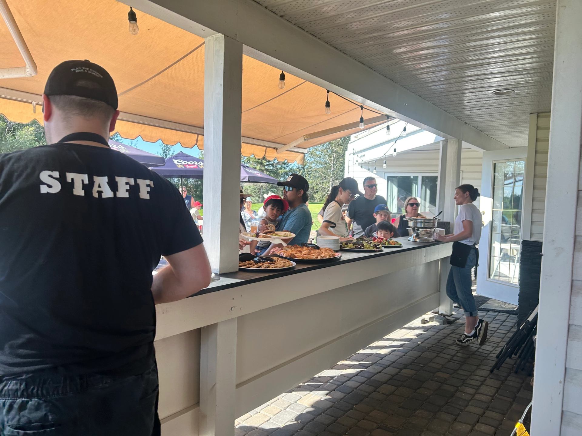 Outdoor serving counter with people eating, under large white tent lights and awnings.