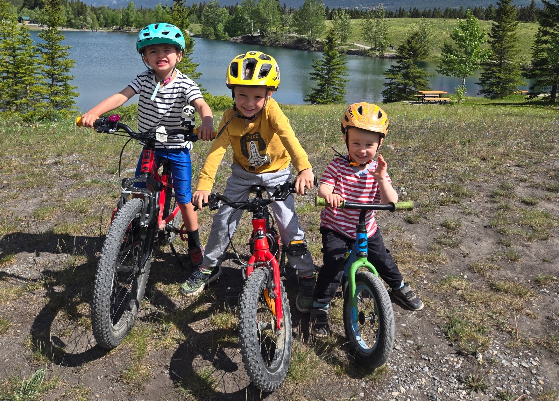 Three children with helmets sit on bikes near a lake and evergreen trees.
