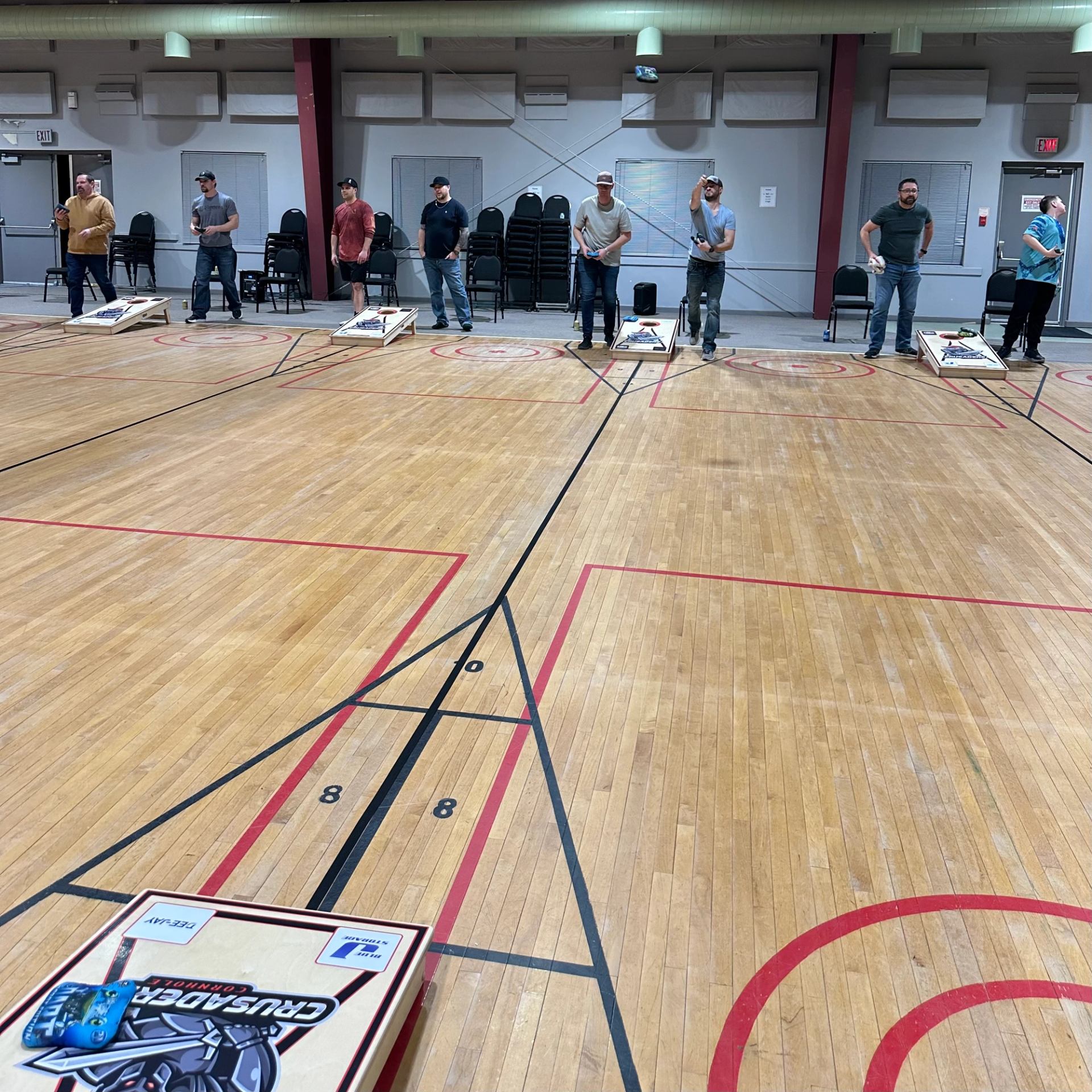 Group of players standing at cornhole boards along a gym floor