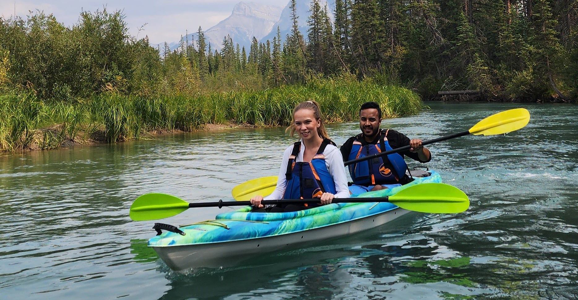 Two kayakers paddle with pine trees and mountains in the background.