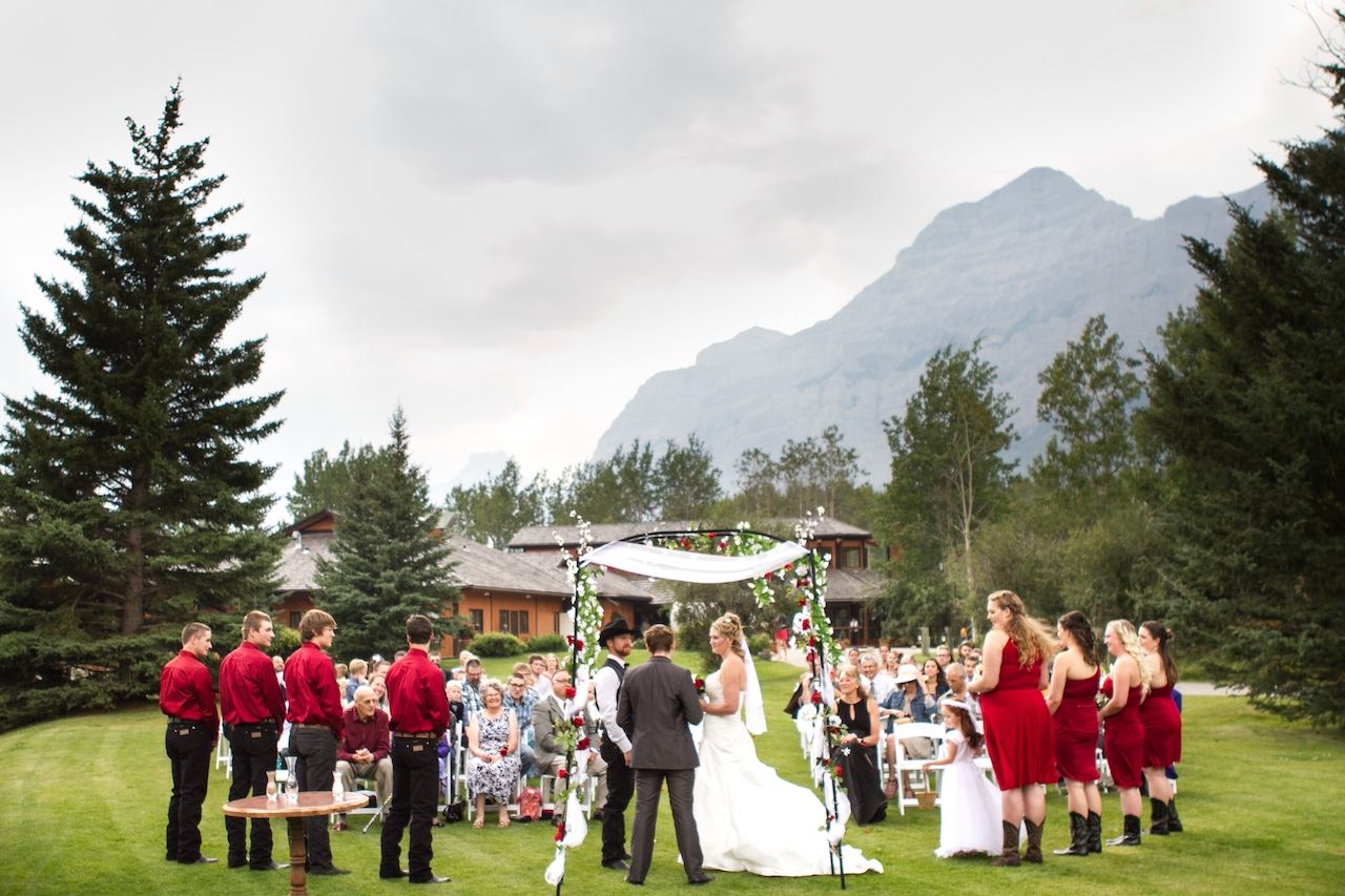 Outdoor wedding ceremony on a green lawn with a floral arch and mountain backdrop.