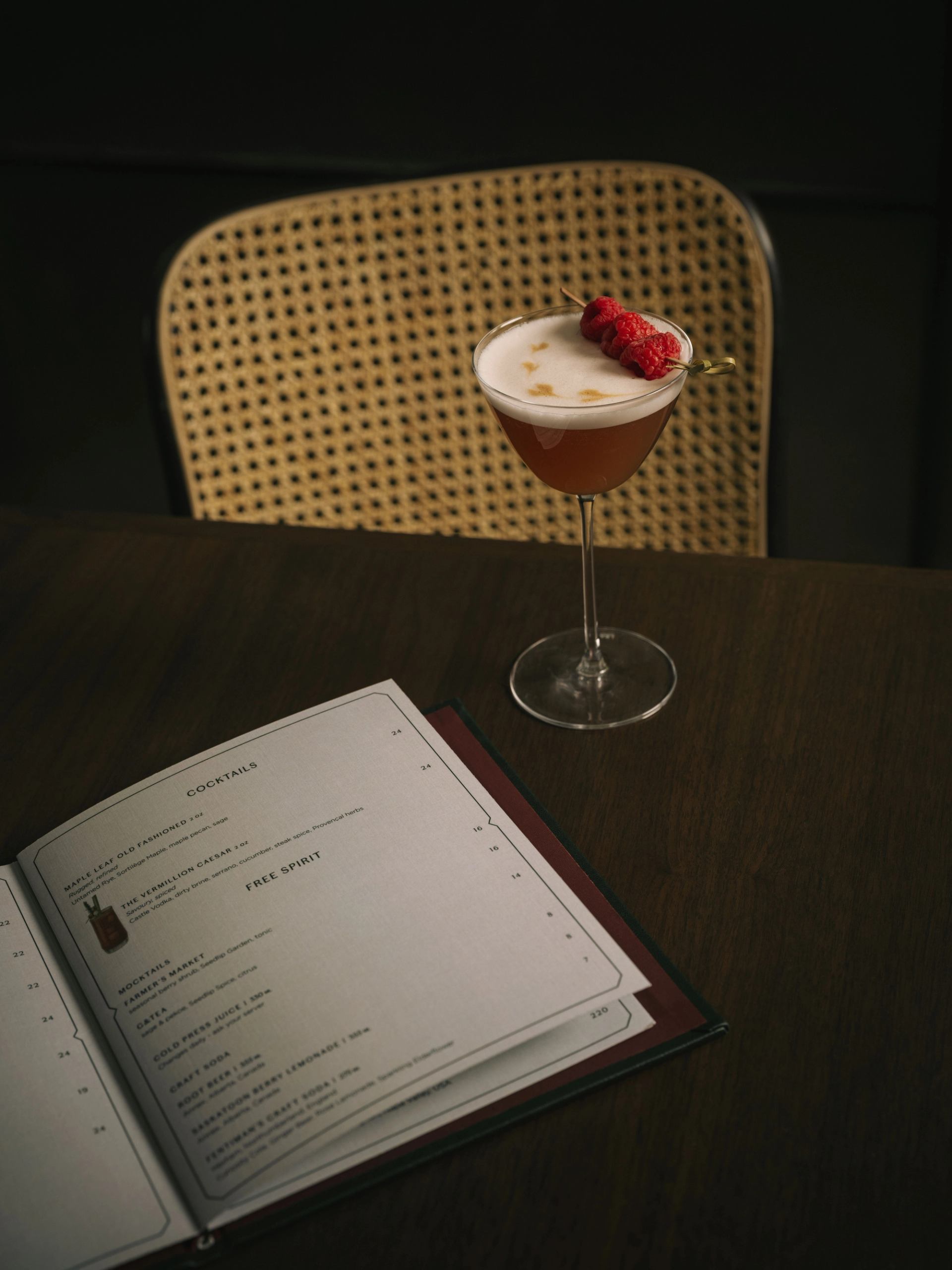 Cocktail with foam and raspberry garnish on a wooden table beside an open drink menu.