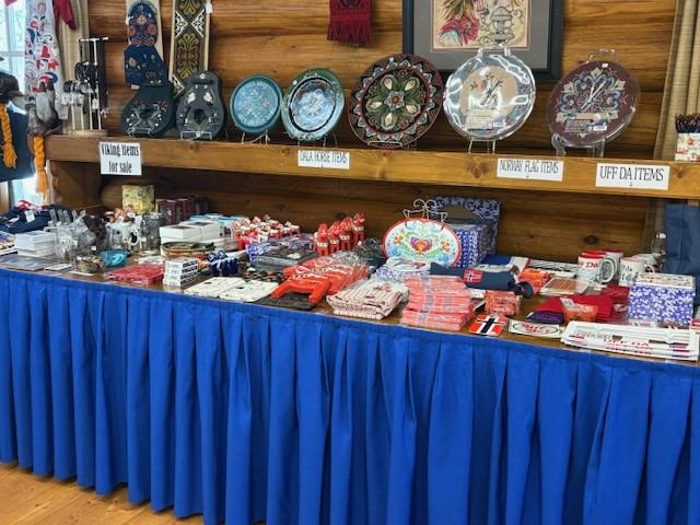 A display table with a blue tablecloth features Norwegian-themed souvenirs and crafts, with decorative items on a wooden shelf above.