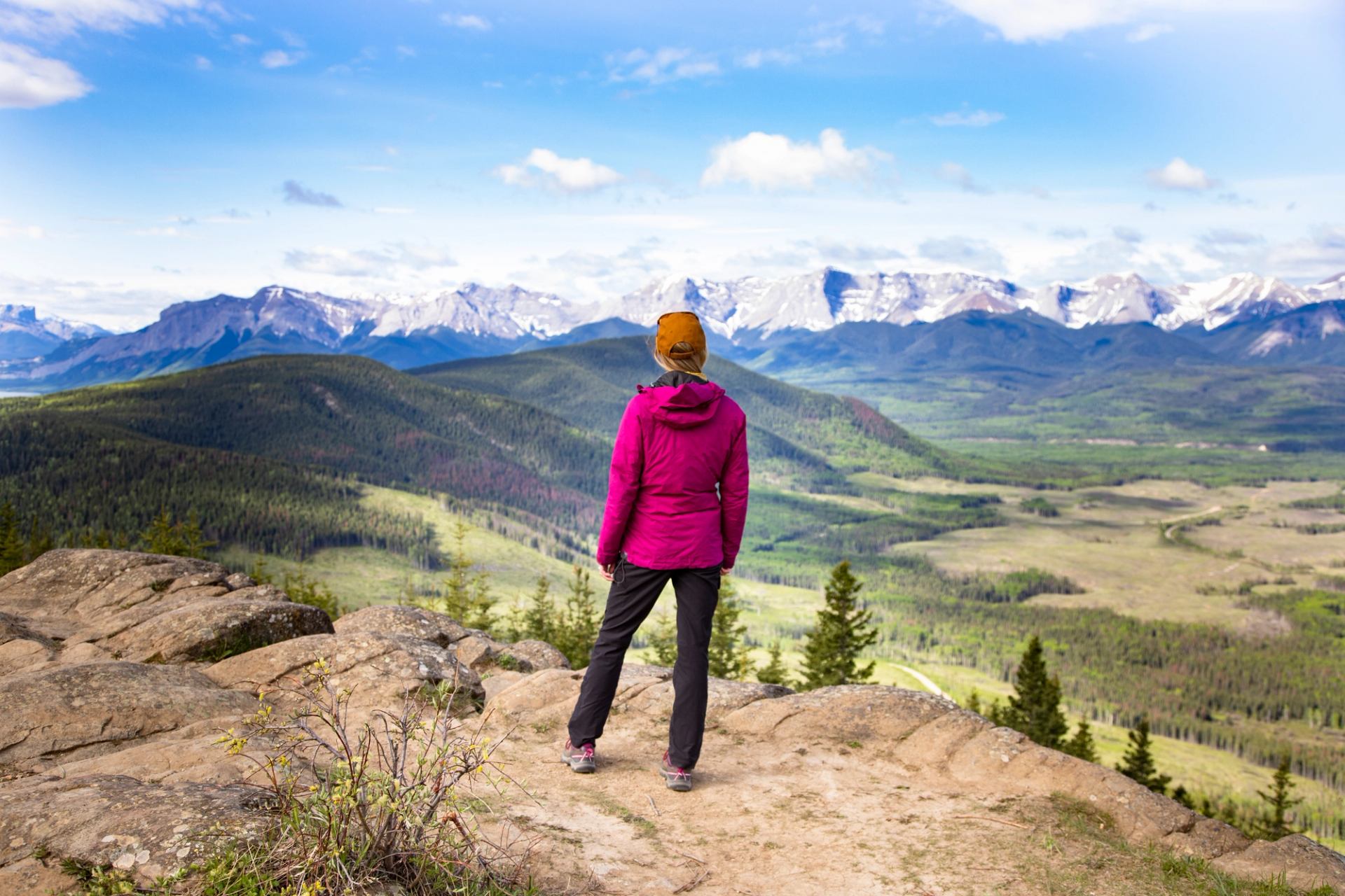 Hiker in pink jacket stands on rocky lookout facing distant snow-capped mountain range.