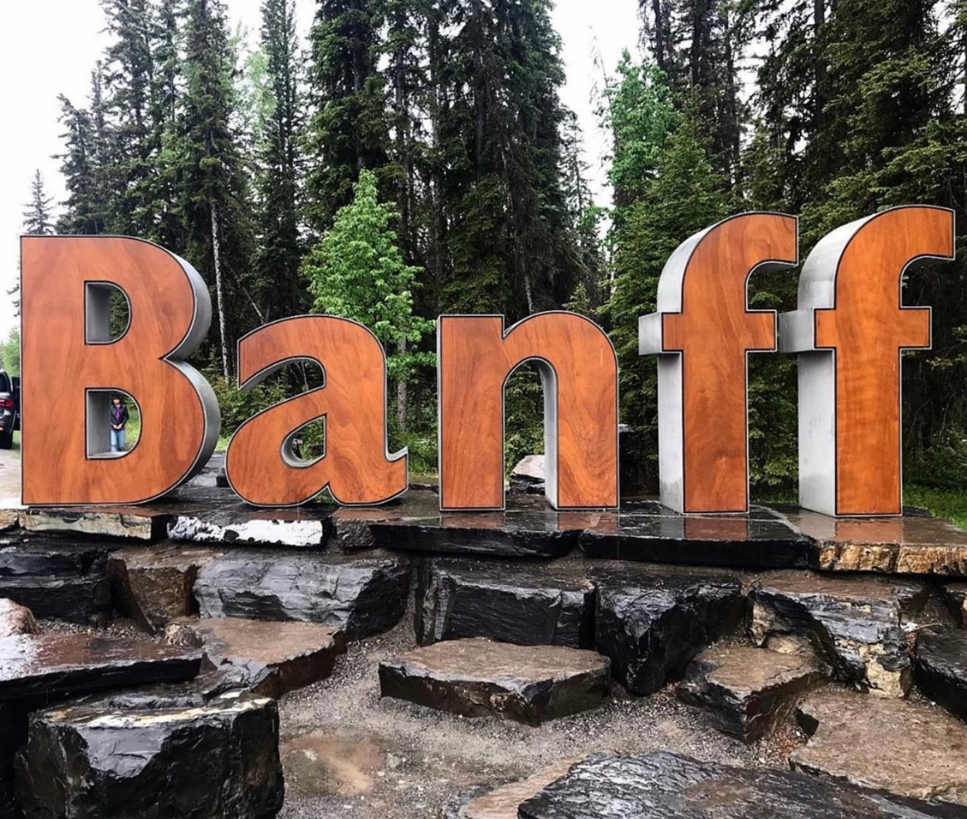 Large Banff sign made of wood and metal, set on rocks with tall pine trees in the background.