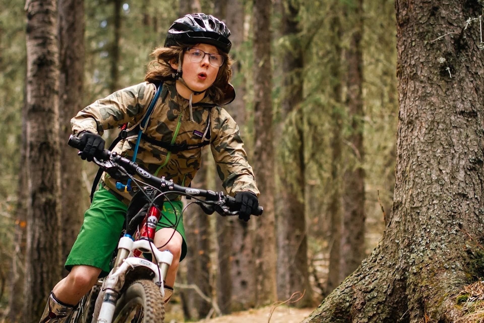Mountain biker wearing helmet and camo jacket rides along forest trail past tall trees.