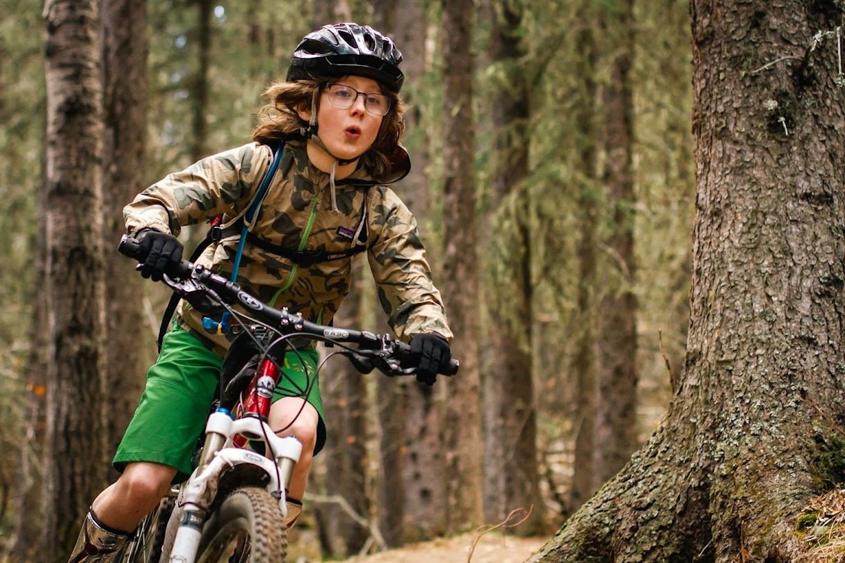 Mountain biker wearing helmet and camo jacket rides along forest trail past tall trees.