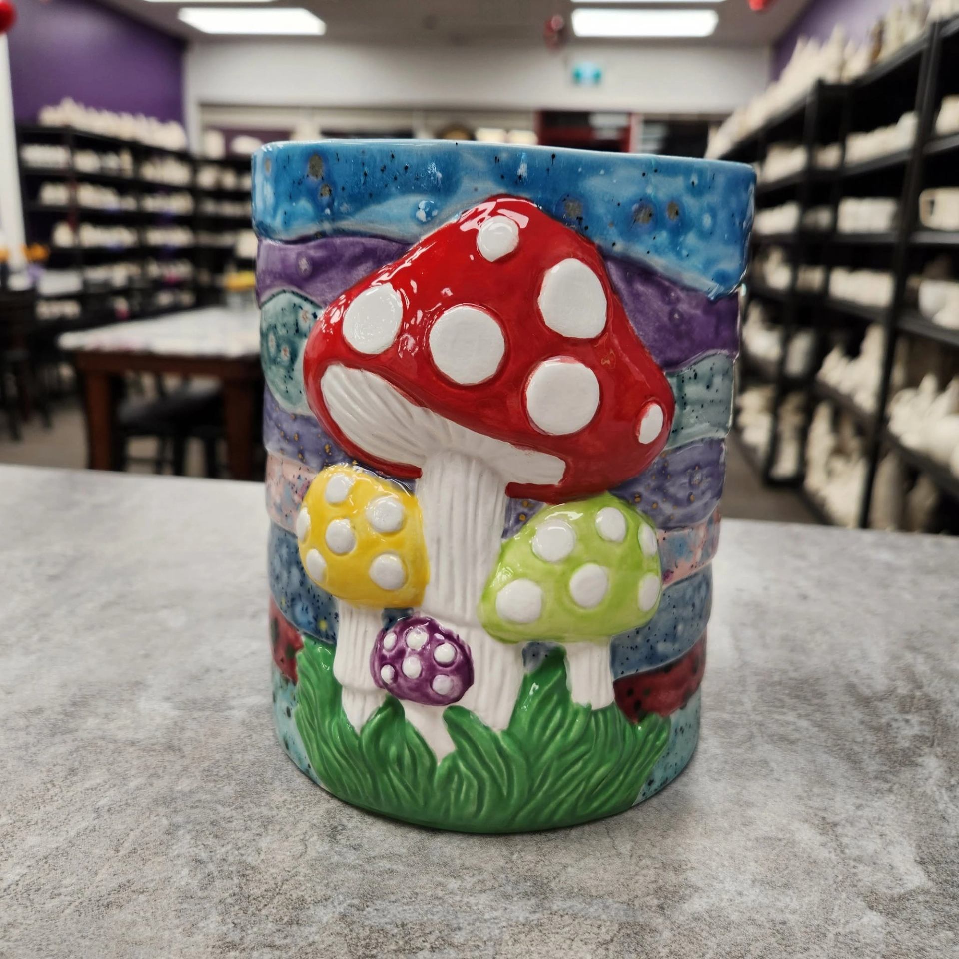 Painted ceramic mug with colourful spotted mushrooms on table in pottery studio.