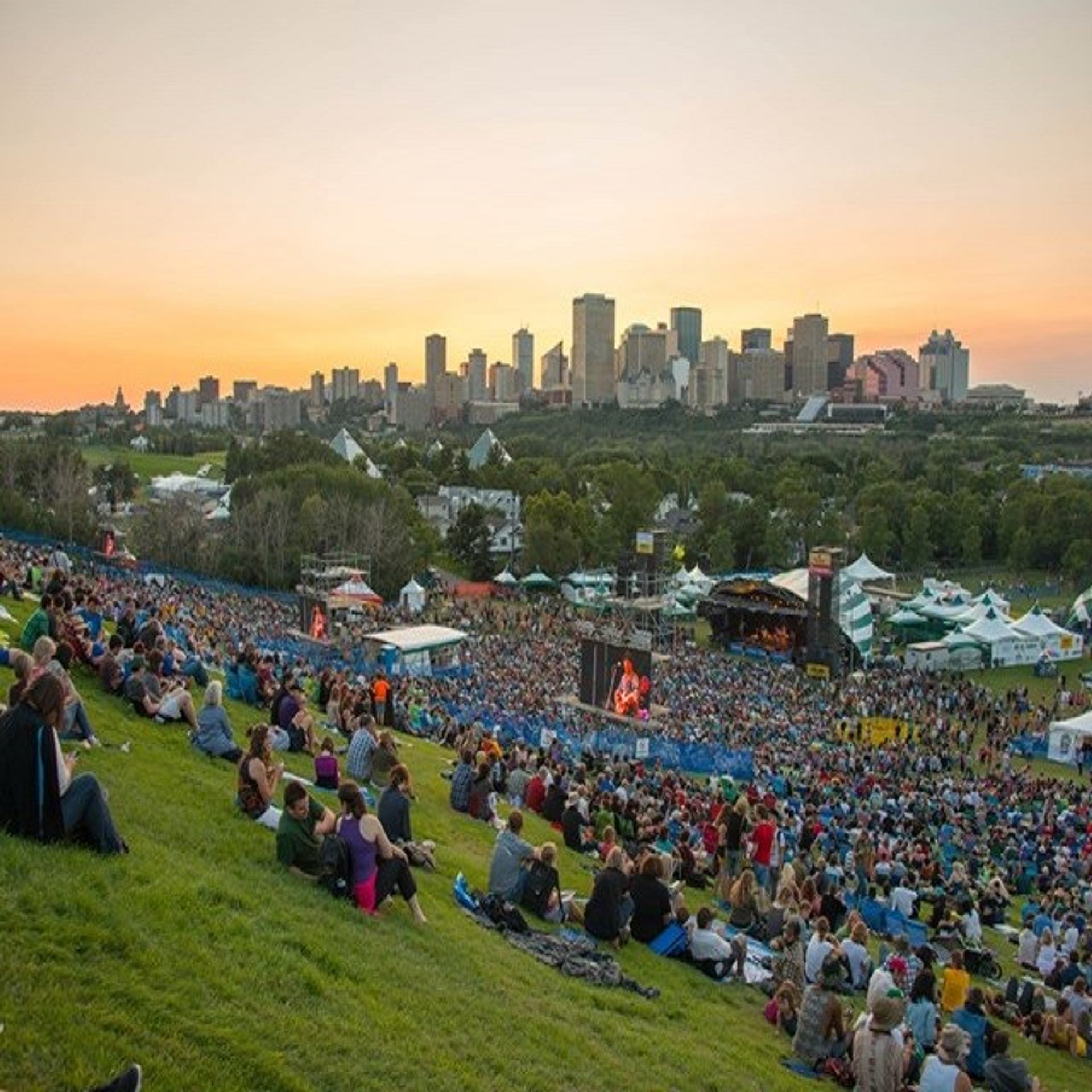 Large crowd on grassy hill watching a stage with Edmonton skyline at sunset