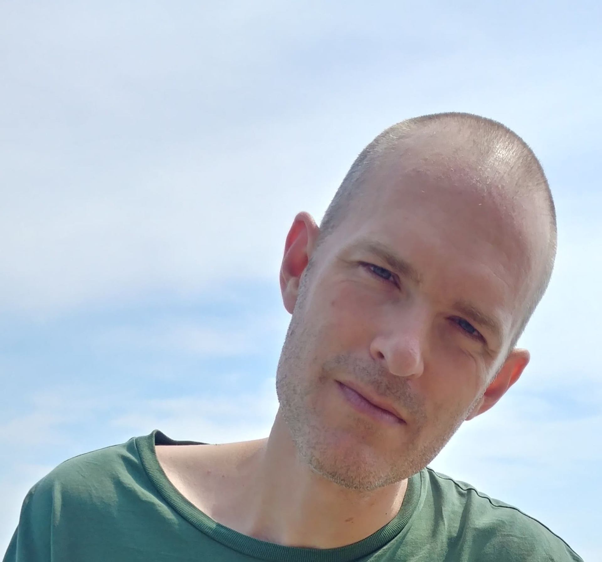 A man with a shaved head and stubble smiles slightly, tilting his head against a blue sky.