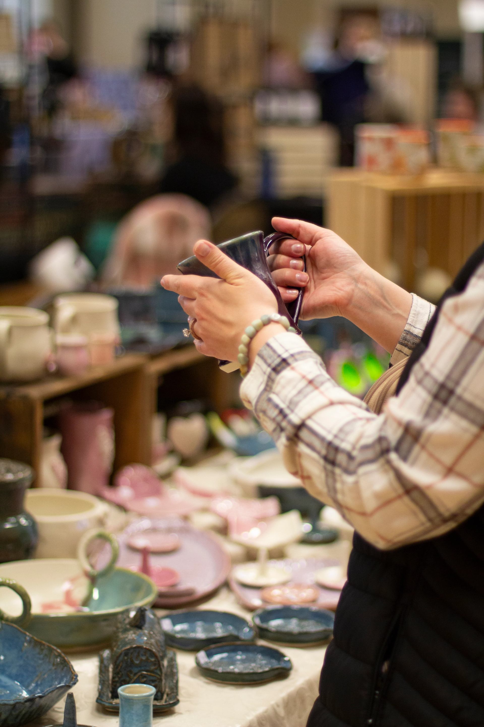 Hands holding a handmade ceramic mug while browsing locally crafted pottery at an indoor market display.