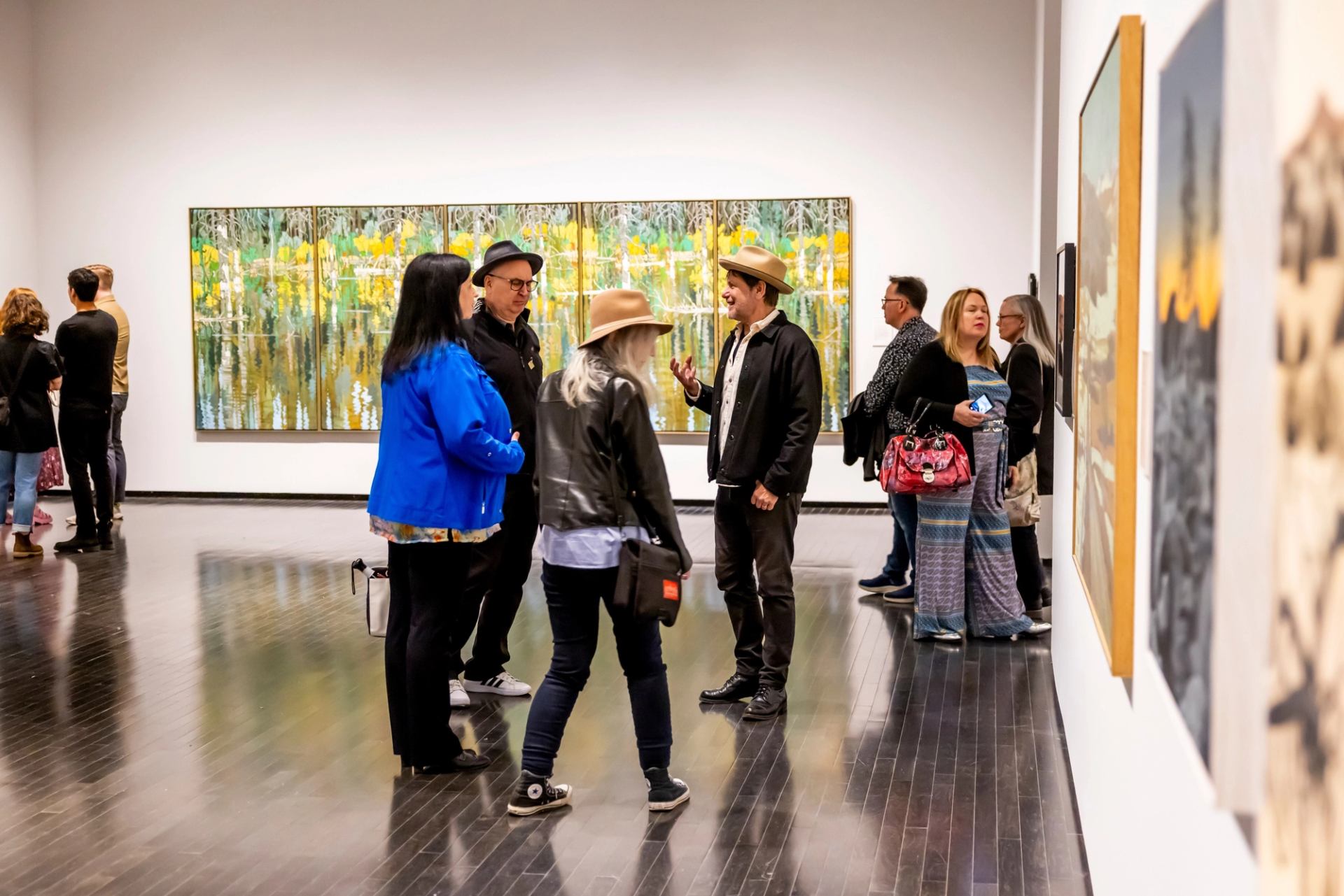 Gallery visitors converse among colourful paintings on white walls.