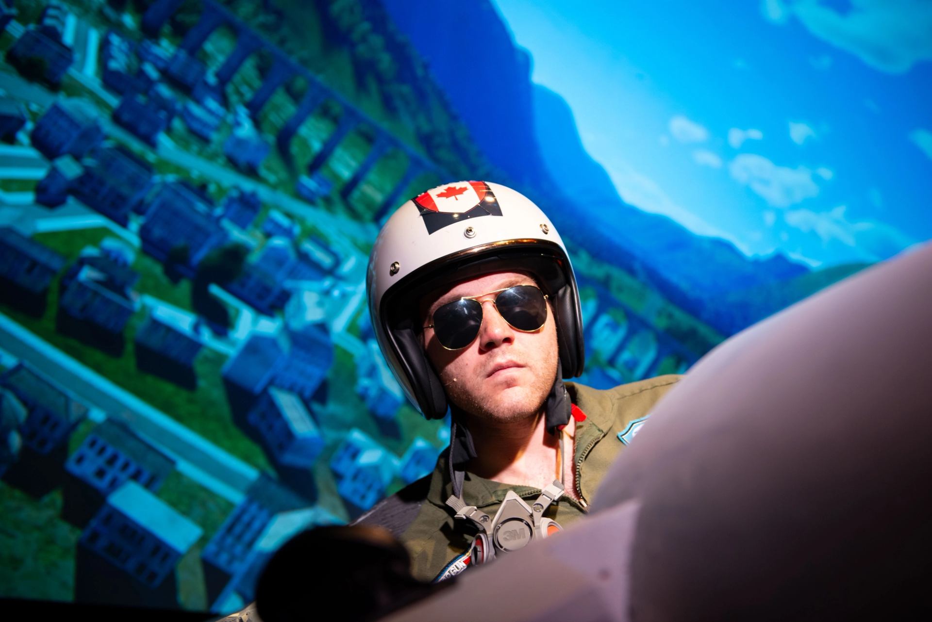 Person wearing a pilot helmet seated in a flight simulator with a cityscape on screen